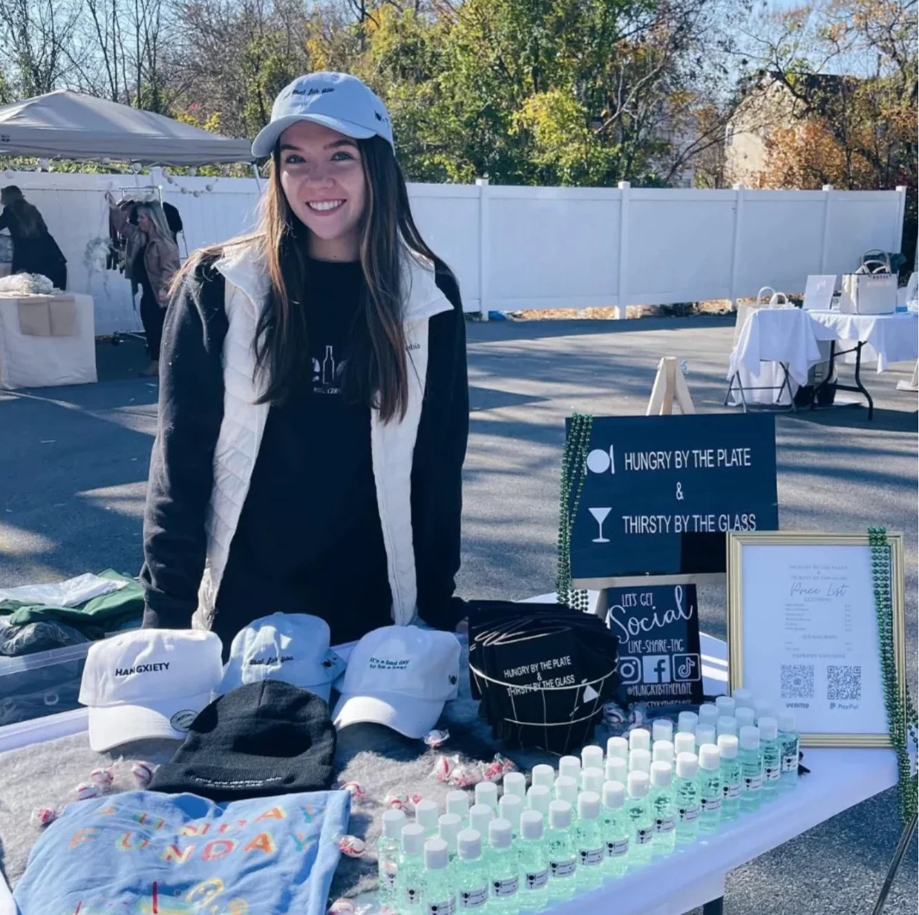 Woman standing behind a table with merchandise and a sign at an outdoor event.